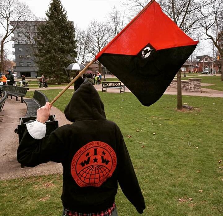 Person in an IWW hoodie (unidentified) holds a black and red flag with Sabo-Tabby on it.