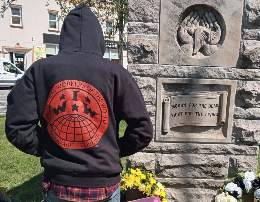 Person in an IWW hoodie (unidentified) stands before a grave for fallen workers.