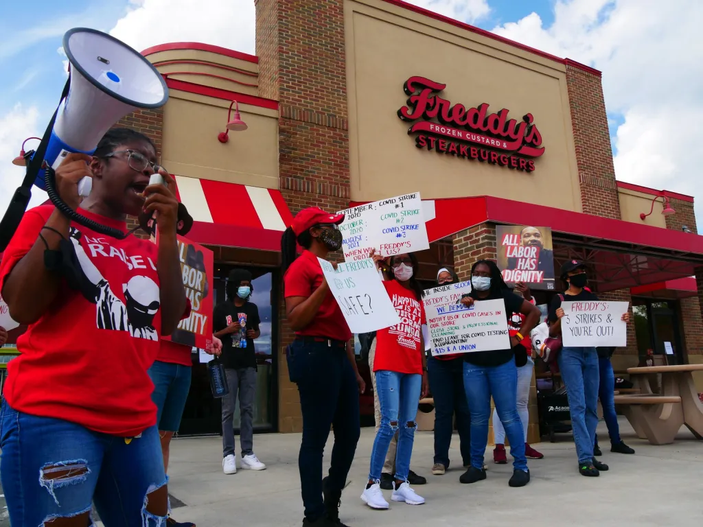 Group of workers on strike in front of a restaurant holding a red cursive sign that reads "Freddys Frozen Custards and Steakburgers." One worker shouts through a megaphone and others hold up signs.