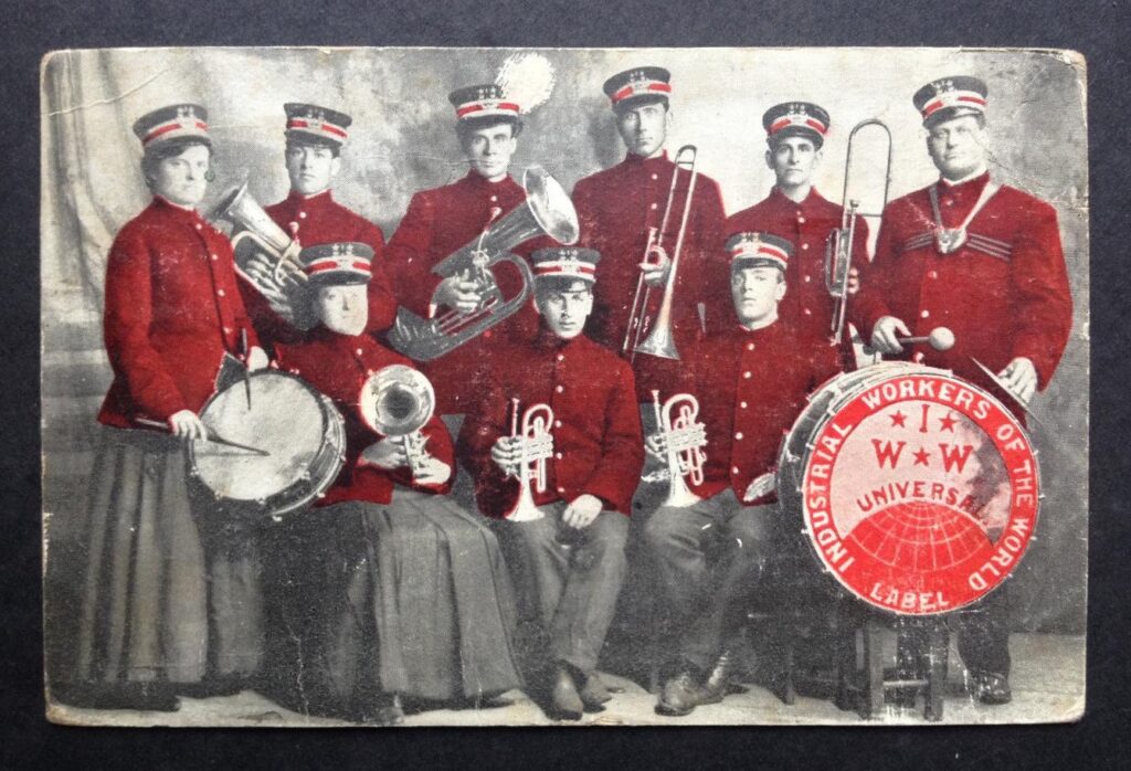 1909 IWW band, nine Wobblies in red marching band uniforms stand together with instruments. IWW Globe logo in corner.