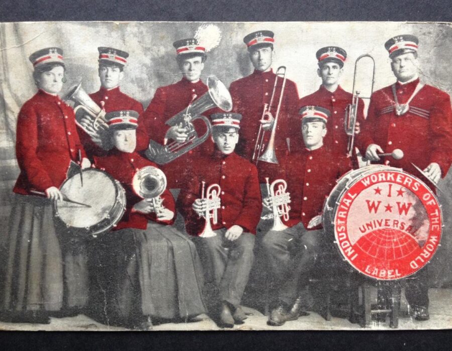 1909 IWW band, nine Wobblies in red marching band uniforms stand together with instruments. IWW Globe logo in corner.