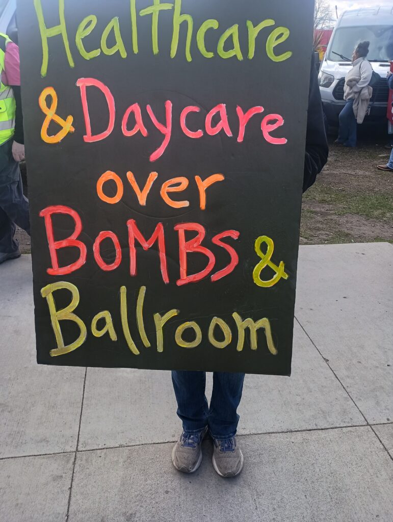 Photo of a person holding a sign that says "Healthcare and Daycare over Bombs and Ballroom."